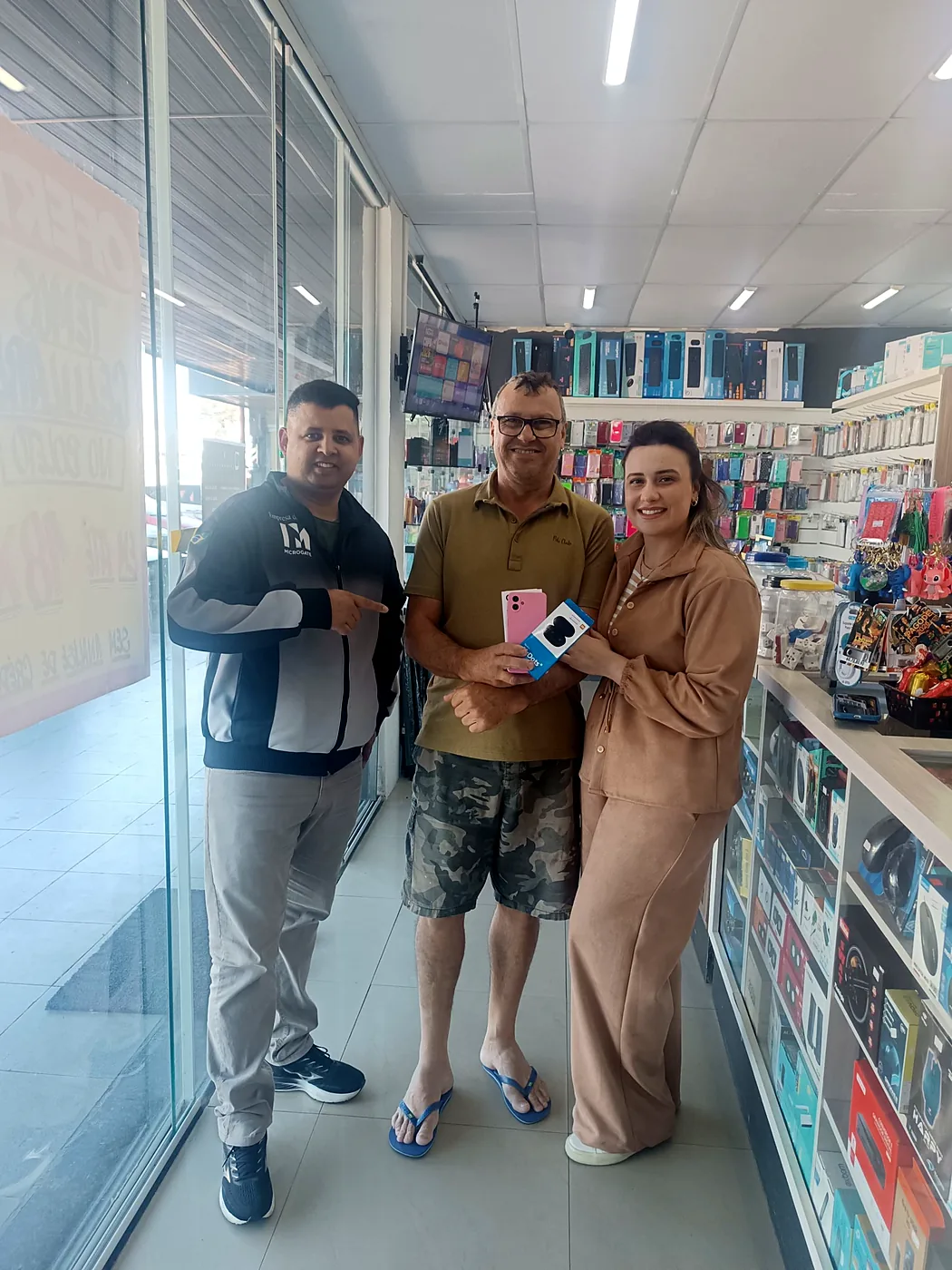 Junior, da Infotech, ao lado de um casal de clientes segurando um celular rosa e a caixa de um fone Redmi