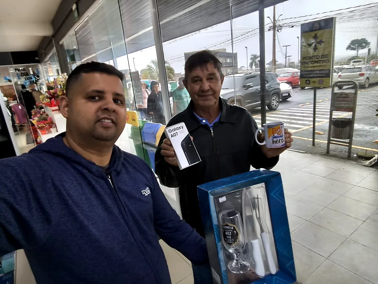 Cliente com a caixa de um Samsung Galaxy A07 na calçada do Boni, ao lado de Junior