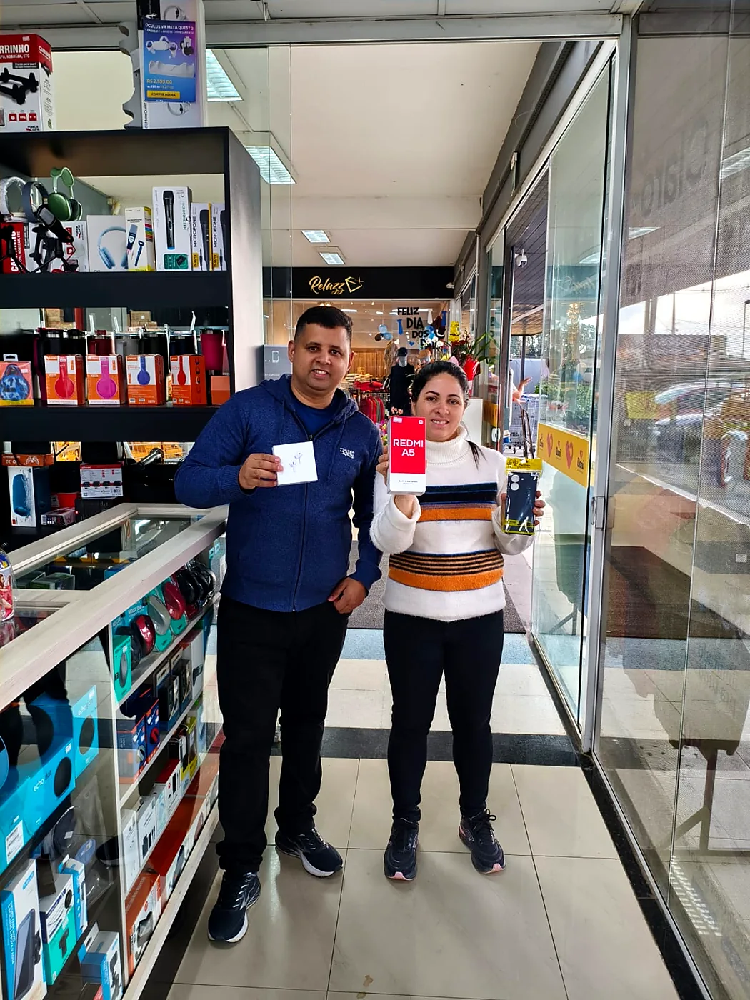Cliente segurando a caixa de um Redmi A5 e uma capa, ao lado de Junior na entrada da loja